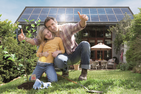 Ein Vater posiert mit seinem Kind stolz der neuen PV-Anlage auf dem Hausdach und zeigt Daumen nach oben.